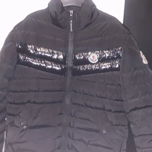 Svart Moncler pufferjacka herr - Svart pufferjacka från Moncler med glansiga paneler över bröstet och klassisk logga på bröstet och ärmen. Jackan har hög krage, dragkedja framtill och två fickor med knappar. Perfekt för kalla dagar och riktigt snygg streetstil.