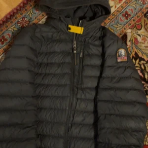 Svart dunjacka Parajumpers L - Svart superlätt dunjacka från Parajumpers med huva, dragkedja och ikonisk patch på ärmen. Jackan har quiltad design och en gul detalj vid halsen. Perfekt för dig som vill ha en snygg och funktionell jacka med streetkänsla.