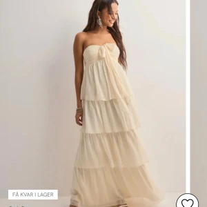 Ljus långklänning från Nelly - Supersöt långklänning från Nelly i ljus cremefärg. Klänningen är axelbandslös med volanger i flera lager och en söt knytdetalj framtill. Perfekt för sommarens alla tillfällen. Luftigt material som ger en drömmig känsla.