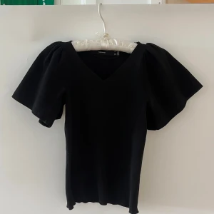 Svart topp med puffärmar från Vero Moda - Svart topp från Vero Moda med v-ringning och stora puffärmar. Modellen är figurnära med korta, vida ärmar som ger en cool siluett. Perfekt för dig som gillar stilrena plagg med en twist. Materialet känns mjukt och stretchigt. Skicka ett meddelande för fler bilder.