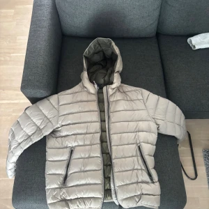 Beige pufferjacka från Colmar - Säljer en beige pufferjacka från Colmar med huva och dragkedja. Jackan har två snedställda fickor med dragkedja och är fodrad i olivgrönt. Perfekt för kyliga dagar och riktigt skön att bära.