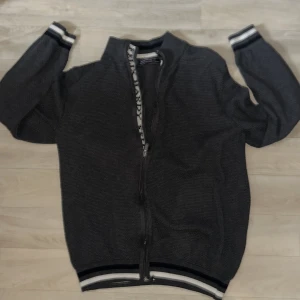 Grå kofta från Felix Hardy  - Mörkgrå kofta från Felix Hardy med hel dragkedja framtill och ribbade muddar med vita och svarta ränder. Storlek L men passar som M mer. Använd få tal gånger/väldigt fräsch