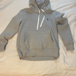 Grå hoodie från Polo Ralph Lauren - Snygg grå hoodie från Polo Ralph Lauren med klassisk blå logga på bröstet. Tröjan har huva med vita snören, känguruficka och ribbade muddar. Perfekt för en chill och stilren look. Den är liten i storleken så rekommenderar att man är runt 178 till 184