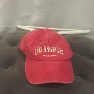 Röd keps Los Angeles vintage - Röd keps i bomull med broderad text 'Los Angeles' framtill och justerbart spänne bak. Klassisk basebollmodell med böjd skärm och avslappnad passform. Perfekt accessoar för en cool streetstyle-look.