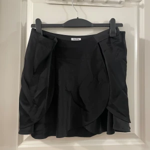 Filippa K svart omlottkjol  - Stilren svart kortkjol från Filippa K med omlottdesign och lätt volangkant. Kjolen har en enkel och elegant look, perfekt för dig som gillar minimalistisk stil. Tillverkad i ett mjukt och följsamt material som ger en snygg passform.