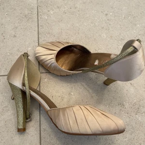 RAVEL Champagnefärgade pumps med glitterklack - Snygga pumps från Pavel i champagnefärgat satin med veckad design framtill. Klacken är täckt av guldglitter och remmen är smal och glittrig. Skorna har spetsig tå och hög klack, perfekta för att sticka ut på speciella tillfällen.