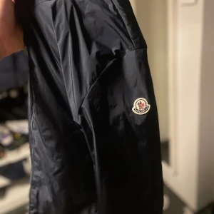 Svart vindjacka från Moncler - Snygg svart vindjacka från Moncler med klassisk logga på ärmen. Jackan har dragkedja framtill och är tillverkad i ett lätt och glansigt syntetmaterial. Perfekt för dig som vill ha en stilren och sportig look.