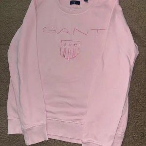 Ljusrosa sweatshirt från Gant - Superfin ljusrosa sweatshirt från Gant med broderad logga och sköld framtill. Tröjan har rund halsringning och långa ärmar, perfekt för en chill och stilren look. Tillverkad i mjuk bomull för extra komfort.