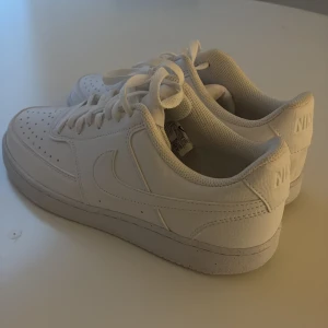 Nike Air Force 1 helvita sneakers - Klassiska Nike Air Force 1 sneakers i helvitt skinn med perforerad tå och vit sula. Snygg och tidlös modell med rund tå, snörning och diskret Nike-logga på sidan och hälen. Perfekta för dig som gillar en clean och stilren look.