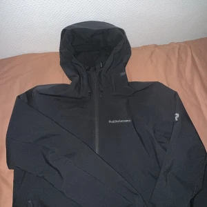 Svart vindjacka Peak Performance - Svart vindjacka från Peak Performance med huva och hel dragkedja framtill. Jackan har diskret logga på bröstet och är tillverkad i ett slätt, syntetiskt material. Justerbar huva med snörning och praktiska fickor med dragkedja.