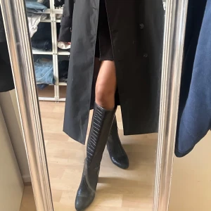 Svarta höga stövlar med klack - Säljer ett par svarta höga stövlar i skinnimitation. Stövlarna har rund tå och stabil hög klack, vilket ger en snygg siluett. Perfekta att styla med kjol eller klänning för en trendig look.