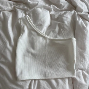 Vit one shoulder ribbad topp - Säljer en stilren vit one shoulder topp i ribbat material. Toppen är ärmlös och har en asymmetrisk skärning som ger en modern vibe. Perfekt att matcha med jeans eller kjol för en clean look. Märke och storlek är lite oklart så lappen skavde så jag klippte bort men passar en xs/s 