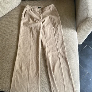 Beiga kostymbyxor från Vero Moda - Stilrena beiga kostymbyxor från Vero Moda med raka ben och pressveck. Byxorna har hällor för bälte och diskreta fickor framtill. Perfekta för en clean och snygg look. Materialet känns mjukt och lätt, vilket gör dem sköna att bära hela dagen.