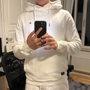 Vit hoodie från Morris, storlek M - Säljer en stilren vit hoodie från Morris i storlek M. Tröjan har huva med snörning, ribbade muddar och en liten broderad logga på bröstet. Tillverkad i mjuk bomull som är skön att ha på sig. Perfekt för dig som gillar en clean och enkel look.