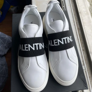 Valentino Open vita sneakers strl 42 - Snygga vita Valentino Open sneakers i skinn med bred svart elastisk rem över foten och tydlig logga. Klassisk rund tå och platt sula. Perfekt för dig som gillar stilrena och exklusiva sneakers med en modern twist. Om du funderar över något skriv så svarar jag, priset är inte hugget i sten vid snabb affär 😊