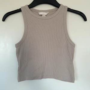 Beige ribbad croppad linnetopp H&M - Säljer en beige croppad linnetopp från H&M. Toppen är ribbad och ärmlös med rund halsringning och tight passform. Perfekt att styla med jeans eller kjol för en clean look.
