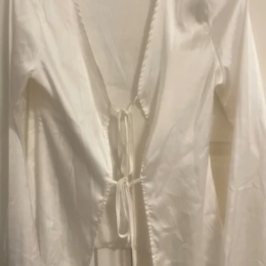 Vit omlottopp med snörning XS - Säljer en vit omlottopp i glansigt material med långa ärmar och snörning framtill. Toppen är tillverkad i 95% polyester och 5% elastan vilket ger en mjuk och följsam känsla. Perfekt att styla med jeans eller kjol för en trendig look.