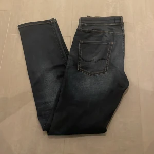 Jack & Jones Slim Straight Tim jeans - Mörkblå jeans från Jack & Jones, modell Slim Straight Tim. Klassisk femficksdesign med snygga slitningar och kontrastsömmar. Jeansen har en rak passform med lite smalare ben och är tillverkade i jeansmaterial. Perfekta för en avslappnad och stilren look.