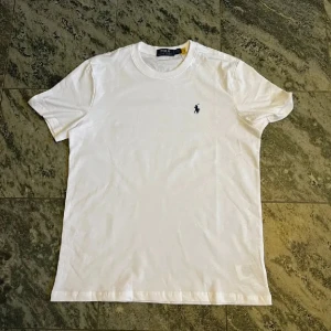 Vit t-shirt från Polo Ralph Lauren - Klassisk vit t-shirt från Polo Ralph Lauren med rund hals och korta ärmar. Liten broderad logga på bröstet. Tillverkad i mjuk bomull för en skön och clean look. Perfekt basplagg till garderoben.