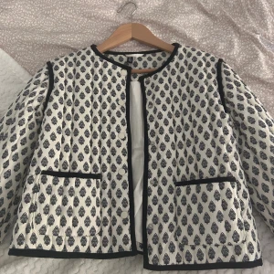 Quiltad mönstrad jacka från H&M Divided - Quiltad jacka från H&M Divided i vitt med svart mönster och svarta kantband. Jackan har rund hals, två fickor framtill och knäppning med tryckknappar. Perfekt för dig som gillar en unik och trendig look.