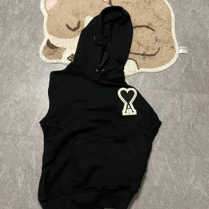 Svart hoodie från Ami Paris med hjärtdetalj - Svart hoodie från Ami Paris med stor vit hjärtdetalj och A-logga på bröstet. Tröjan har huva med snörning, känguruficka och långa ärmar. Tillverkad i mjuk bomull för en skön och avslappnad look.