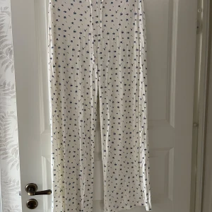 Vita byxor med blåbärsmönster - Supermjuka och lätta vita byxor med blått blåbärsmönster över hela tyget. Byxorna har rak passform och resår i midjan för extra komfort. Perfekta för chill dagar hemma eller som mysbyxor. Materialet känns svalt och luftigt.