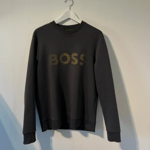 Sweatshirt från Hugo Boss  - Mörkgrå sweatshirt från BOSS med guldig logga framtill. Perfekt för en stilren och avslappnad look. Andvänt skick enligt bilder