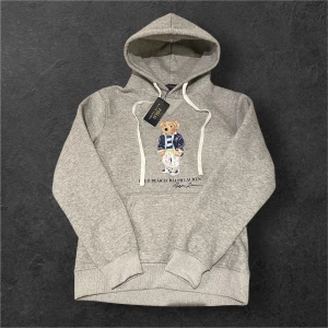 Grå hoodie Polo Bear Ralph Lauren  - Grå hoodie från Polo Ralph Lauren med ikoniska Polo Bear-trycket framtill. Tröjan har huva med vita snören och en stor magficka. Tillverkad i mjuk bomullsmix, perfekt för chill dagar. Klassisk och stilren design med snygga detaljer.