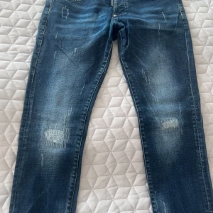 Dsquared2 blå jeans med slitningar - Snygga blå jeans från Dsquared2 med coola slitningar och patchdetaljer bak. Klassisk femficksmodell med raka ben och normal passform. Jeansen har en tydlig Dsquared2-logga och är tillverkade i robust denim för en trendig look.