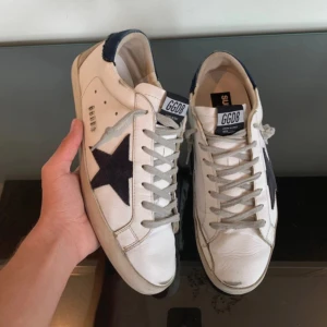 Golden goose skor  - Snygga vita skor från Golden Goose med svart stjärna på sidan och GGDB-logga på plösen. Skorna har grå snörning, svarta detaljer vid hälen och är tillverkade i skinn. Perfekta för dig som gillar en trendig och avslappnad stil. Inget og tillkommer 