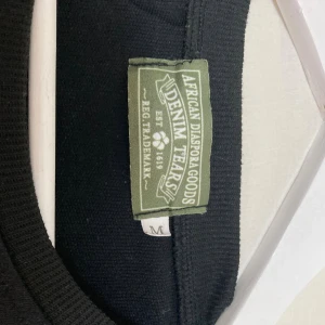 Svart tröja från Denim Tears med tryck - Svart tröja från Denim Tears med unika vita blommönster tryckta över hela plagget. Tröjan har rund halsringning och är tillverkad i mjuk bomull, perfekt för dig som vill sticka ut med en cool och modern stil.