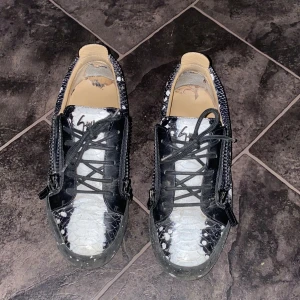Svarta och vita sneakers från Giuseppe Zanotti - Unika sneakers från Giuseppe Zanotti med svart och vit reptilmönstrad ovandel, snörning och dragkedjor på sidorna. Skorna har en platt sula och rund tå, samt coola detaljer som signatur-logga på plösen. Perfekta för dig som vill sticka ut med din stil. / Pris kan diskuteras! / Kan även utbytas mot något.