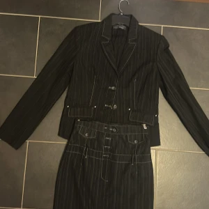 Svart randig kostym med kjol - Säljer en svart kostym med smala vita ränder. Setet består av en figursydd kavaj med knappar, fickdetaljer och markerade sömmar samt en matchande pennkjol med hög midja, knappar och fickor framtill. Klassisk och stilren look med coola kontrastsömmar. Behöver inte köpas som set kontakta om du vill ha endast kavajen/kjolen. 