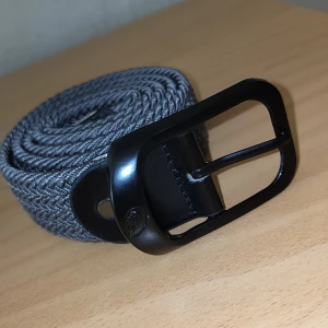 Grått flätat bälte med svart spänne - Snyggt grått flätat bälte från Callaway med svart metallspänne och detaljer i svart skinn. Bältet har en stilren look och passar perfekt till jeans eller chinos. Flätad design gör det enkelt att justera längden för bästa passform.