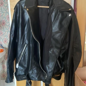 Svart skinnjacka från Zara XXL - Säljer en svart skinnjacka från Zara i storlek XXL. Jackan har klassisk bikerstil med sned dragkedja, stora slag och bälte nedtill. Flera fickor med dragkedja och coola detaljer i silver. Perfekt för dig som vill ha en edgy look.