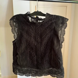 Svart spetsblus från ONLY - Supersnygg svart blus från ONLY med detaljerad spets och vackra broderier. Blusen är ärmlös med vågformade kanter och rund halsringning. Perfekt att styla med jeans eller kjol för en trendig look.