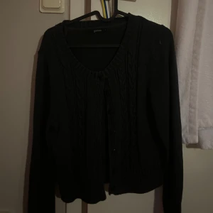 grå stickad kofta från Gianni - grå stickad kofta från Gianni med snyggt flätmönster framtill och v-ringad hals. Koftan har långa ärmar, ribbade muddar och knäppning med svarta knappar. Perfekt för lager-på-lager och en stilren look.