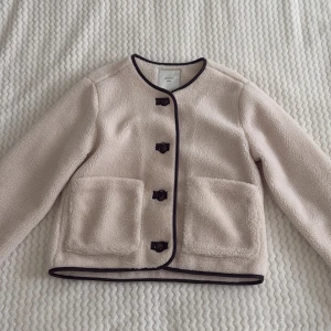 Beige teddyjacka från Neo Noir - Säljer en beige teddyjacka från Neo Noir med svarta kantdetaljer och stora framfickor. Jackan har rund hals, knäppning med svarta knappar och är långärmad. Jättemysig men har tyvärr inte kommit tills användning 