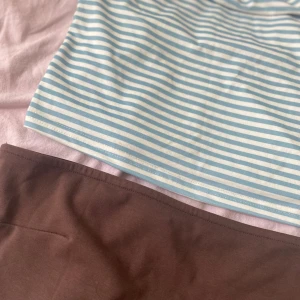 Randig blåvit och brun tubtopp - Säljer två bandeau tubtoppar, en med ljusblå och vit randigt mönster och en i enkel brun färg. Båda är ärmlösa och har en croppad passform, perfekta för varma dagar. Materialet känns mjukt och stretchigt, troligen bomull eller syntet.