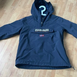 Mörkblå vindjacka från Napapijri helt ny - Snygg mörkblå vindjacka från Napapijri med huva och halv dragkedja. Jackan har en stor ficka framtill med logotyp och norsk flagga, samt långa ärmar. Perfekt för dig som gillar streetwear och vill ha en stilren look.