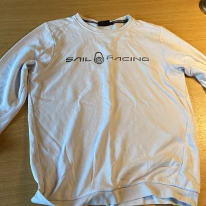 Vit långärmad tröja från Sail Racing - Vit långärmad tröja från Sail Racing med tryckt logga på bröstet. Tröjan har rund halsringning och ribbade muddar vid ärmslut och nederkant. Perfekt för en clean och sportig stil. Tillverkad i mjuk bomull som känns skön mot huden.