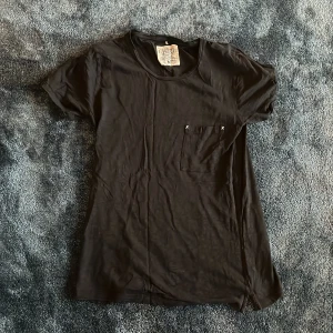 Svart t-shirt från Ljung med ficka - Svart t-shirt från Ljung i storlek M. Modellen har rund halsringning och en bröstficka med två vita korsstygn som detalj. T-shirten är enkel och stilren, tillverkad i mjuk bomull med korta ärmar och normal passform.