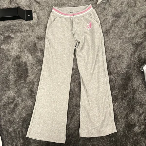 Grå mjukisbyxor från abercrombie and fitch med rosa detalj - Grå mjukisbyxor med breda ben och elastisk midja med rosa och vit rand. Byxorna har fickor och en broderad rosa bokstav på ena höften. Perfekta för chill dagar eller när du vill vara extra bekväm. S/XS