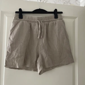 Beige shorts i bomull med snörning - Säljer ett par ljusbeige shorts i mjuk, krinklad bomull med elastisk midja och snörning framtill. De har en avslappnad passform och är perfekta för varma dagar. Shortsen har fickor i sidorna och är riktigt sköna att bära.