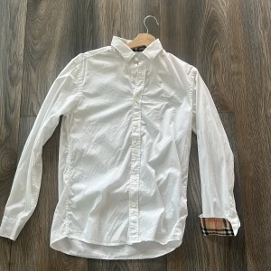 Vit Burberry skjorta med rutiga detaljer - Säljer en klassisk vit skjorta från Burberry i bomull. Skjortan har långa ärmar, knappar framtill och ikoniska rutiga detaljer på insidan av kragen och ärmsluten. Perfekt för dig som gillar stilrena plagg med en twist. Diskret broderad logga på bröstet. Köpt på nk kvitto finns och tags