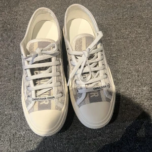 Christian Dior grå sneakers  - Snygga sneakers från Christian Dior i grått och vitt med tydlig Dior-logga på ovandelen och snörning. Skorna har rund tå, platt sula och är tillverkade i textil med gummidetaljer. Perfekta för dig som vill ha en exklusiv och trendig look. Nypris runt 9000kr!! Säljer billigt eftersom vill bli av med allt. I storlek 41 men passar 38-39,5!