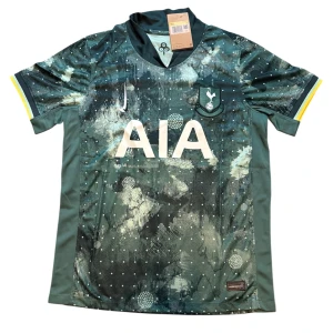 Tottenham Hotspur bortatröja Nike - Snygg Tottenham Hotspur bortatröja från Nike med unikt abstrakt mönster i mörkblått och grönt. Tröjan har klubbmärke på bröstet, vit AIA-logga och gula detaljer på ärmsluten. Tillverkad i lätt och ventilerande polyester, perfekt för match eller träning.