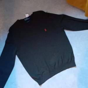 Grön v-ringad tröja från Polo Ralph Lauren - Klassisk grön v-ringad tröja från Polo Ralph Lauren i 100% bomull. Tröjan har lång ärm och den ikoniska röda loggan broderad på bröstet. Perfekt för dig som gillar stilrena och tidlösa plagg.