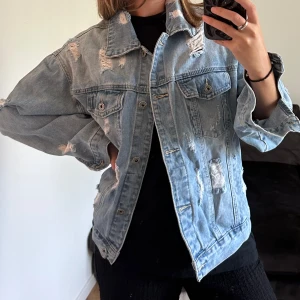 Oversized jeansjacka från Newplay - Säljer en ljusblå oversized jeansjacka från Newplay med slitna detaljer och klassisk krage. Jackan har två bröstfickor med knappar och långa ärmar. Perfekt för dig som gillar en chill och edgy stil.
