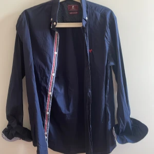 Mörkblå skjorta från Williot - Snygg mörkblå skjorta från Williot med regular fit. Skjortan har vita knappar, diskret röd logga på bröstet och coola detaljer med randigt band längs knappslån. Insidan av kragen har prickigt mönster och ärmsluten har rutigt tyg. Perfekt för dig som gillar stilrena detaljer.